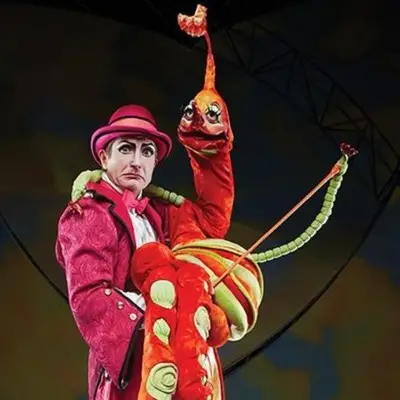 Moha Samedi a performer from Cirque du Soleil's Mystère, dressed in a pink suit and top hat, holding a large, elongated red and orange puppet