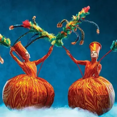 Two performers from Cirque du Soleil's Mystère, wearing large, orange and red striped bell-shaped skirts and conical hats, holding feathery, colorful props.