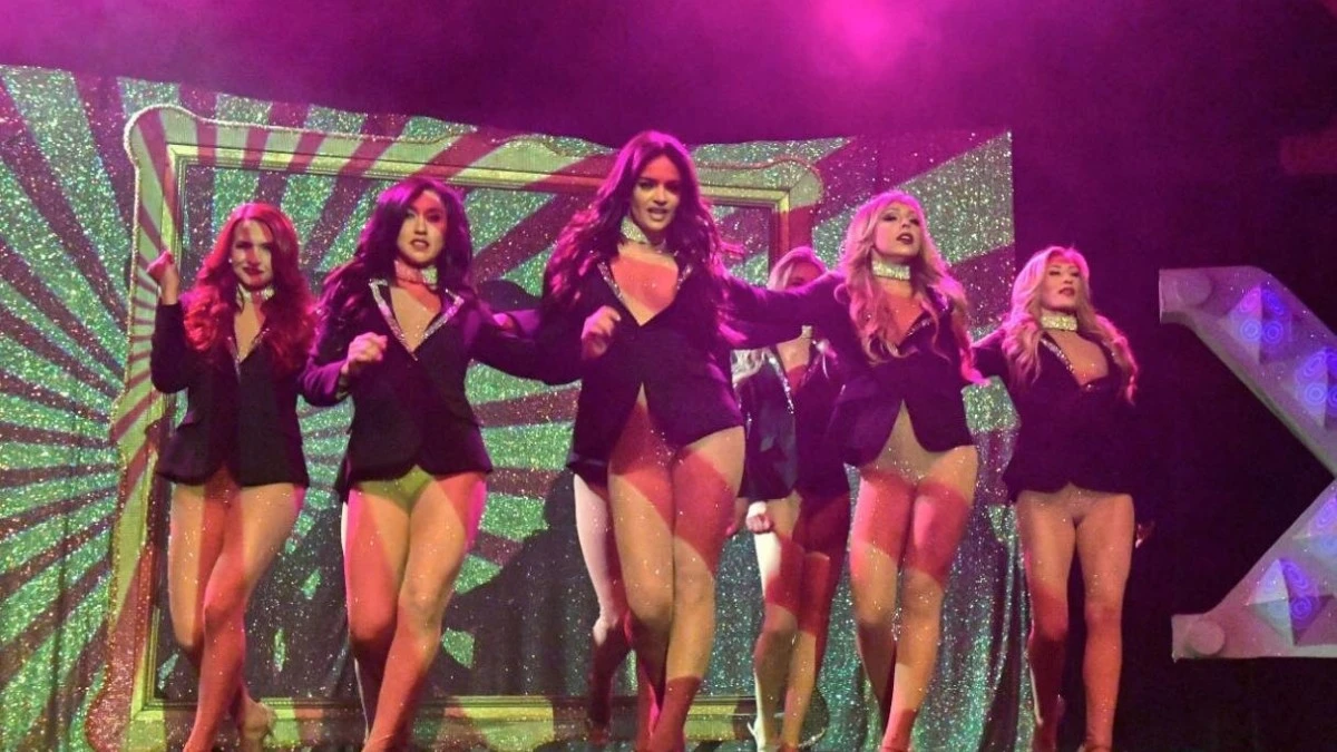 Burlesque performers dancing on stage in sparkling outfits and blazers under colorful stage lights.