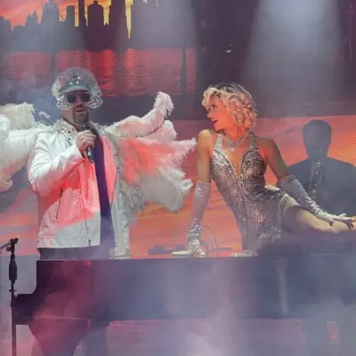 Piano Man performing in white feathered costume and a female singer in sparkling dress.