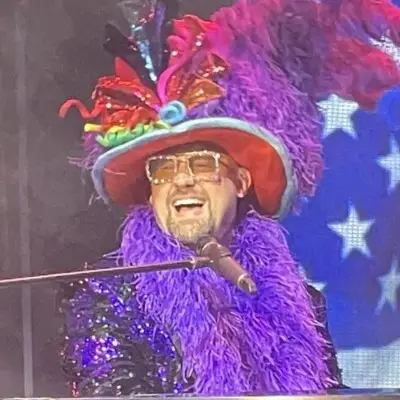 Piano Man performer smiling widely wearing a colorful feathered hat and sequined jacket during a live show.