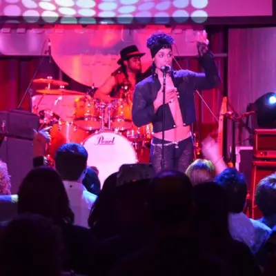 Purple Reign performs on stage in front of an audience with a drummer in the background.