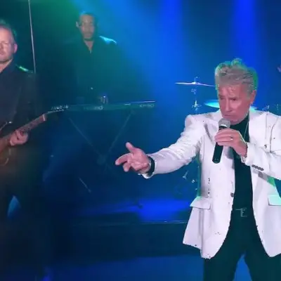 Valli look-alike pointing at the audience while other members are playing instruments
