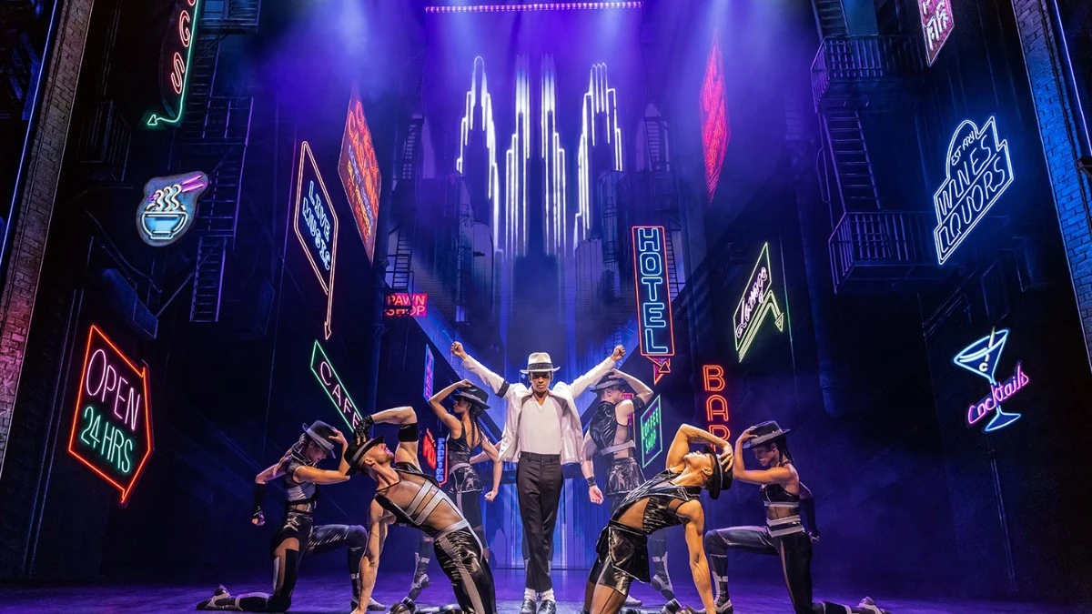 Performer dressed as Michael Jackson mid-dance move on stage with dynamic red lighting and backup dancers in the background during a scene from MJ: The Musical.